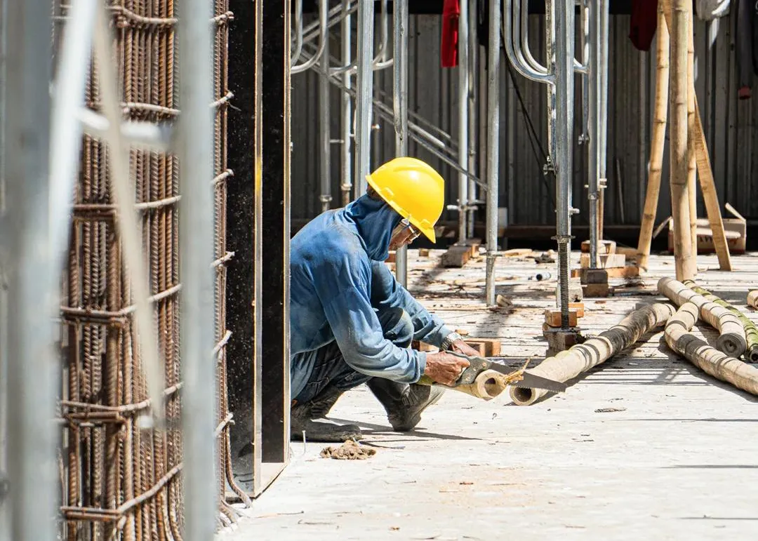 A builder in a yellow hard hat crouches on a concrete slab, cutting bamboo beside steel rebar.