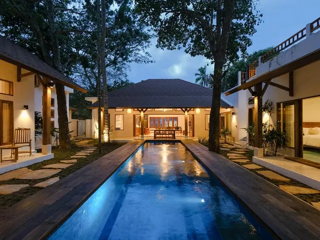 An aerial view of a resort: a long roof over a pool deck with parasols and loungers, lawns and palms around it.