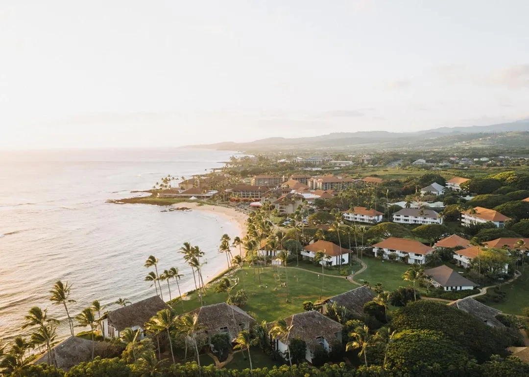 An aerial view at sunset of a palm-lined beach resort, with tiled roofs running back from the sea.