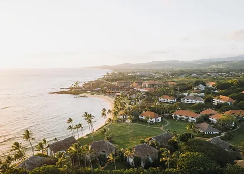 An aerial view at sunset of a palm-lined beach resort, with tiled roofs running back from the sea.