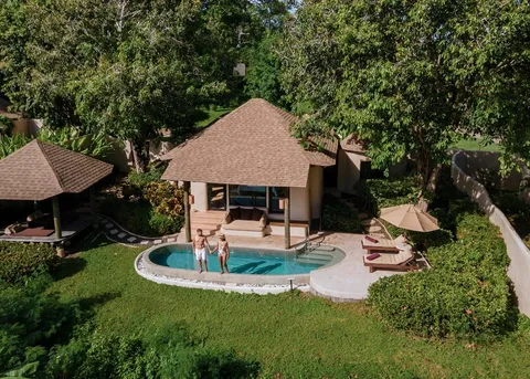An aerial view of a thatched villa with a private pool in a tropical garden, a couple standing at the water's edge.