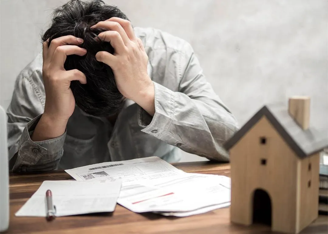 A man holds his head over a pile of papers on a desk, beside a small wooden model house.