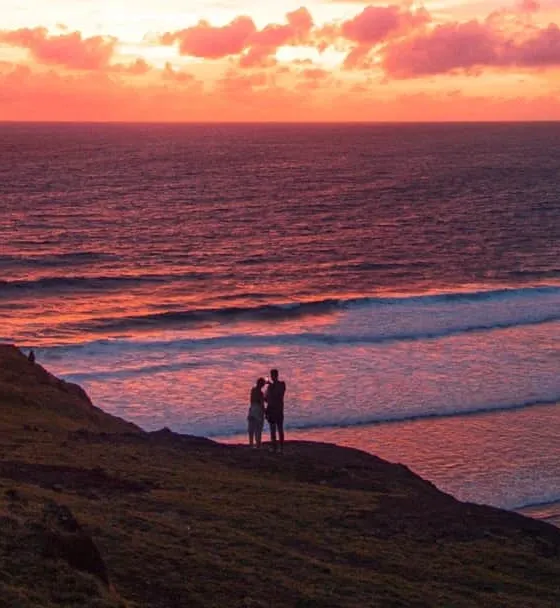 Two people standing on a clifftop above the south Lombok coast at sunset, with waves rolling in below.