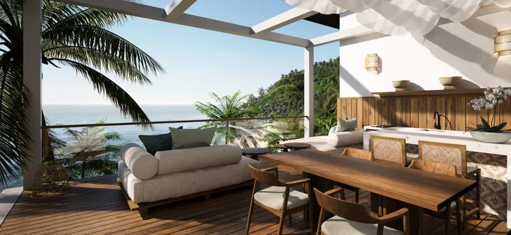 A render of a two-bedroom villa's deck: a white sofa and a long timber dining table under a pergola, with palms and the sea past the glass rail.